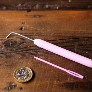 Puede incluir: Un gancho de ganchillo rosa y una aguja de lana rosa sobre una superficie de madera. Tambi&eacute;n se puede ver una moneda de oro en la imagen.