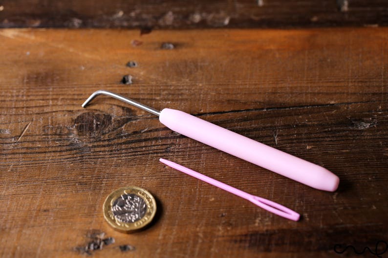Puede incluir: Un gancho de ganchillo rosa y una aguja de lana rosa sobre una superficie de madera. Tambi&eacute;n se puede ver una moneda de oro en la imagen.