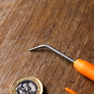 Puede incluir: Un gancho de ganchillo peque&ntilde;o de mango naranja con un gancho de metal plateado. El gancho est&aacute; sobre una superficie de madera junto a una moneda.