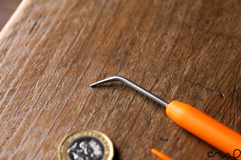 Puede incluir: Un gancho de ganchillo peque&ntilde;o de mango naranja con un gancho de metal plateado. El gancho est&aacute; sobre una superficie de madera junto a una moneda.