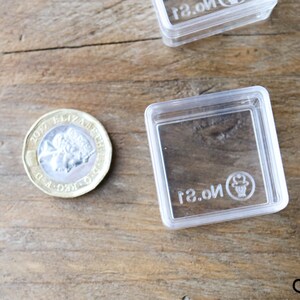 May include: A close-up of a British one pound coin with the inscription "2017 ELIZABETH II D.G. REG. F.D."  The coin is lying on a wooden surface next to a clear plastic square box with the text "No. 21" printed on the bottom.