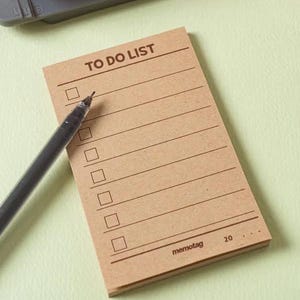 May include: A rectangular tan paper notepad with the text TO DO LIST printed at the top, featuring seven horizontal lines with square checkboxes on the left side. A black ballpoint pen rests diagonally across the notepad, and a grey hole puncher is visible.