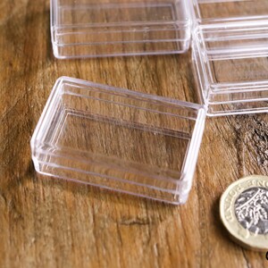 May include: Clear plastic rectangular hinged boxes, ideal for small item storage, are displayed on a wooden surface. A British one-pound coin is also present, providing a size reference. The boxes are empty and ready for use.