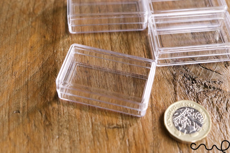 May include: Clear plastic rectangular hinged boxes, ideal for small item storage, are displayed on a wooden surface. A British one-pound coin is also present, providing a size reference. The boxes are empty and ready for use.
