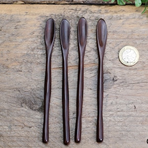 Puede incluir: Cuatro cucharas de madera de color marrón oscuro con extremos redondeados. Las cucharas están dispuestas en fila sobre una superficie de madera.