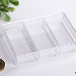 May include: A clear, rectangular plastic organizer with three compartments. The organizer is empty and sits on a white surface. A gold coin is next to the organizer. The organizer is designed for small items.