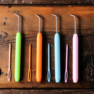 Puede incluir: Seis ganchillos de crochet de colores con puntas met&aacute;licas, dispuestos en fila sobre una superficie de madera. Los ganchillos son verde, naranja, azul, rosa y dos son melocot&oacute;n.