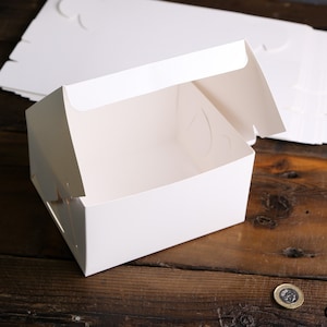 Könnte beinhalten: Eine weiße, offene Bäckerei-Box mit gefaltetem Deckel, auf einer dunklen Holzoberfläche. Mehrere zusätzliche Boxen sind im Hintergrund gestapelt. Die Box hat eine rechteckige Form mit kleinen Griffen an den Seiten.