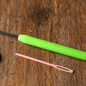 Puede incluir: Una aguja de lana verde y una aguja de tapicer&iacute;a rosa sobre una superficie de madera.