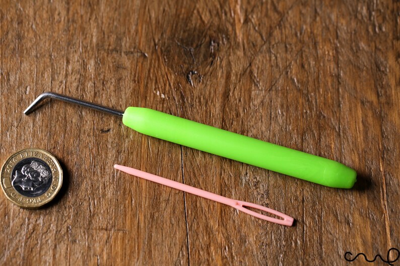 Puede incluir: Una aguja de lana verde y una aguja de tapicer&iacute;a rosa sobre una superficie de madera.