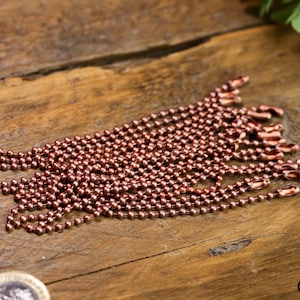 Puede incluir: Un montón de cadena de bola de cobre, con cada cadena terminando en un pequeño bucle. Las cadenas están dispuestas en forma de abanico sobre una superficie de madera.