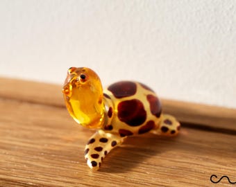 以下が含まれることがあります： 半透明の琥珀色の頭と首にダークブラウンの斑点がある小さなガラス製のカメの置物。甲羅はクリームイエローで、大きな不規則なダークブラウンの斑点があり、ヒレは黄色でダークブラウンの斑点模様が施されています。