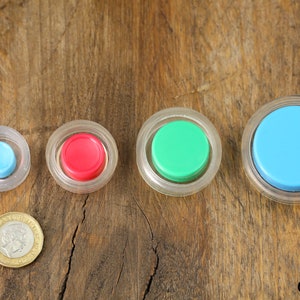 May include: Four round push buttons in different sizes. The buttons are encased in clear plastic and have coloured centres: blue, red, green, and light blue.