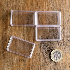 May include: Clear rectangular plastic coin holders, some open and some closed, are arranged on a wooden surface. A gold-colored coin with a detailed design is also present. The holders are designed to protect and display coins.