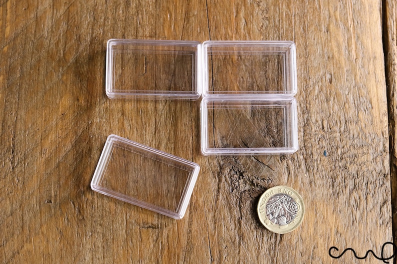 May include: Clear rectangular plastic coin holders, some open and some closed, are arranged on a wooden surface. A gold-colored coin with a detailed design is also present. The holders are designed to protect and display coins.