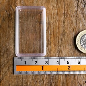 May include: A clear plastic coin holder next to a British one-pound coin and a ruler. The ruler shows measurements in inches. The coin is gold and silver in color and has text on it.