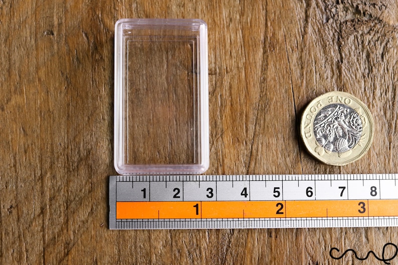 May include: A clear plastic coin holder next to a British one-pound coin and a ruler. The ruler shows measurements in inches. The coin is gold and silver in color and has text on it.