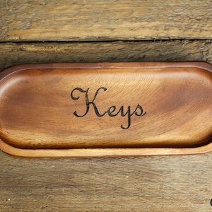 以下が含まれることがあります： 前面に「Keys」と刻まれた木製のキーホルダー。トレーは楕円形で、自然な木製の仕上げです。