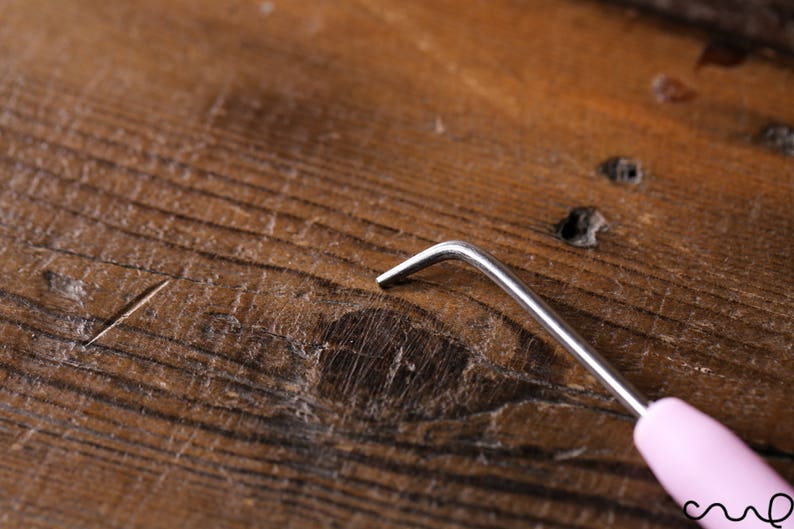 Puede incluir: Primer plano de un gancho de ganchillo rosa con un gancho de metal plateado. El gancho est&aacute; sobre una superficie de madera.