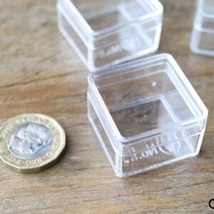 May include: Four clear plastic square boxes with lids. The boxes are stacked on top of each other. A British one pound coin is in the foreground.