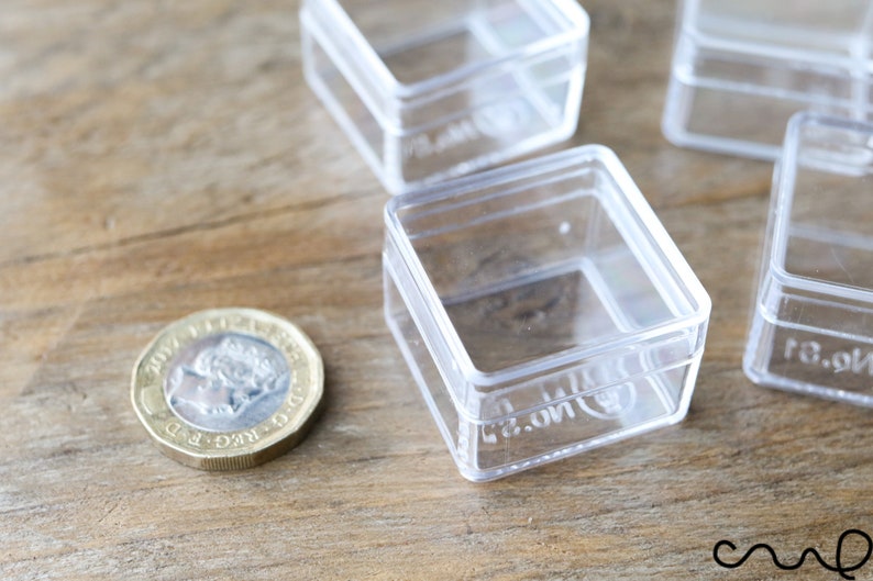 May include: Four clear plastic square boxes with lids. The boxes are stacked on top of each other. A British one pound coin is in the foreground.