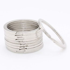 May include: A stack of silver-colored key rings with a split ring design. One key ring is separated from the stack, showing the open split. The key rings are made of metal and are on a white background.
