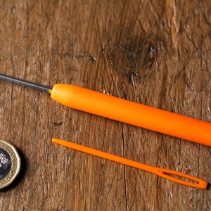Op de afbeelding: Twee oranje garennaalden op een houten oppervlak. De grotere naald heeft een gebogen metalen punt en een plastic handvat. De kleinere naald is recht en heeft een klein oog aan het ene uiteinde. Een Britse munt van &eacute;&eacute;n pond is ook zichtbaar op de afbeelding.