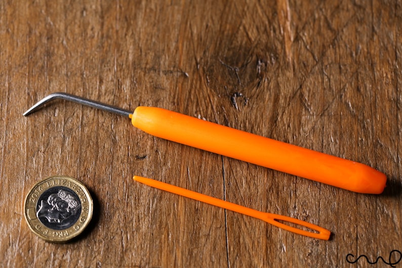 Op de afbeelding: Twee oranje garennaalden op een houten oppervlak. De grotere naald heeft een gebogen metalen punt en een plastic handvat. De kleinere naald is recht en heeft een klein oog aan het ene uiteinde. Een Britse munt van &eacute;&eacute;n pond is ook zichtbaar op de afbeelding.
