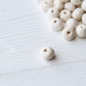 May include: A close-up of a single white ceramic bead with a hole in the center. The bead is sitting on a white wooden surface. There are other white ceramic beads scattered in the background.