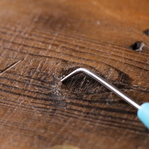 Op de afbeelding: Een close-up van een houten oppervlak met een klein, metalen haakgereedschap met een blauwe handgreep. Het gereedschap wordt gebruikt om kleine voorwerpen uit krappe ruimtes te verwijderen.
