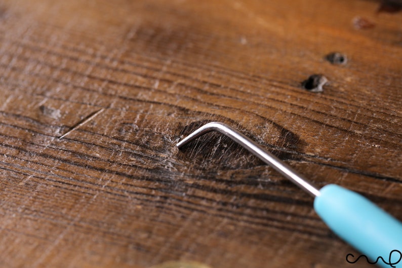 Op de afbeelding: Een close-up van een houten oppervlak met een klein, metalen haakgereedschap met een blauwe handgreep. Het gereedschap wordt gebruikt om kleine voorwerpen uit krappe ruimtes te verwijderen.