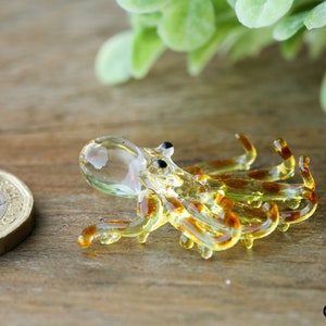 Pu&ograve; includere: Una piccola statuina di polpo in vetro con un corpo trasparente e strisce marroni e gialle. Il polpo &egrave; sdraiato di lato con i tentacoli arrotolati.