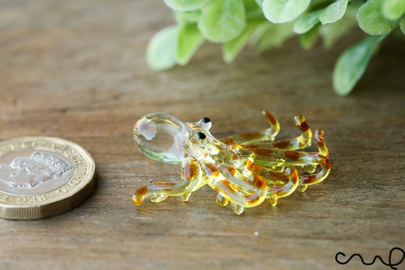 Pu&ograve; includere: Una piccola statuina di polpo in vetro con un corpo trasparente e strisce marroni e gialle. Il polpo &egrave; sdraiato di lato con i tentacoli arrotolati.