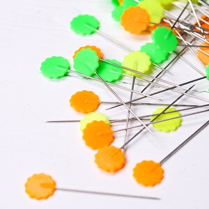 May include: A close-up of a pile of colorful sewing pins with green, orange, and yellow plastic heads. The pins are scattered on a white surface.