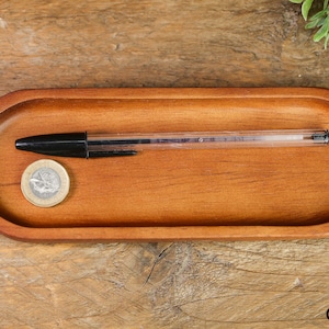 Puede incluir: Una bandeja ovalada de madera con un bolígrafo negro y una moneda de plata en su interior. La bandeja está hecha de madera marrón claro y tiene un acabado liso.