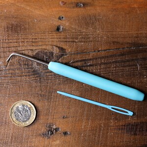 Puede incluir: Un gancho de crochet de pl&aacute;stico azul y una aguja de lana de pl&aacute;stico azul sobre una superficie de madera. Tambi&eacute;n se puede ver una moneda brit&aacute;nica de una libra.