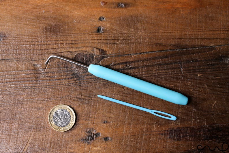 Puede incluir: Un gancho de crochet de pl&aacute;stico azul y una aguja de lana de pl&aacute;stico azul sobre una superficie de madera. Tambi&eacute;n se puede ver una moneda brit&aacute;nica de una libra.