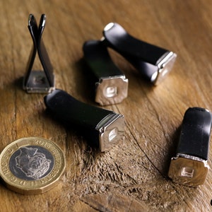 May include: A collection of black and silver metal clips, possibly for holding papers or other small items, are arranged on a wooden surface. A British one-pound coin is also present. The clips have a rectangular base and a curved, black top.