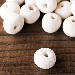 Puede incluir: Un montón de cuentas de cerámica blanca con un agujero en el centro de cada cuenta. Las cuentas están sobre una superficie de madera.