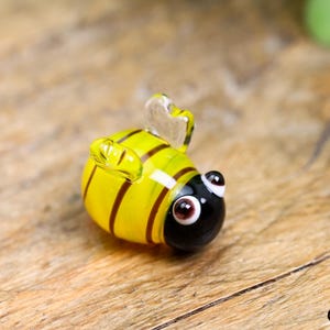 May include: A small, glass bee figurine. The bee has a yellow body with dark brown stripes, a black head, and clear wings. The eyes are brown with white highlights. The figurine is resting on a wooden surface.