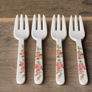 May include: Four white forks with floral rose designs. The forks have three tines and a handle decorated with pink roses, blue accents, and green leaves. The forks are arranged on a wooden surface.