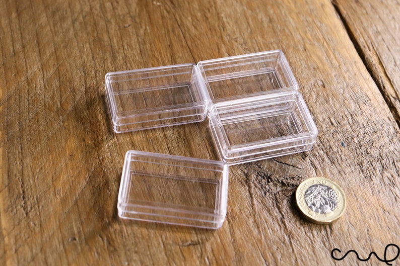 May include: Four clear, rectangular plastic boxes are arranged on a wooden surface. A British one-pound coin is next to the boxes. The boxes appear to be empty and are likely for small items.