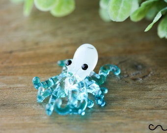 May include: A small glass octopus figurine with a white body and blue tentacles. The octopus is sitting on a wooden surface.