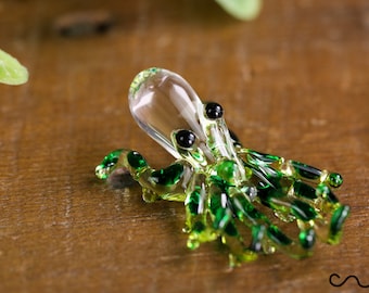 May include: A small glass octopus figurine with a clear body and green tentacles. The octopus has black eyes and is sitting on a wooden surface.