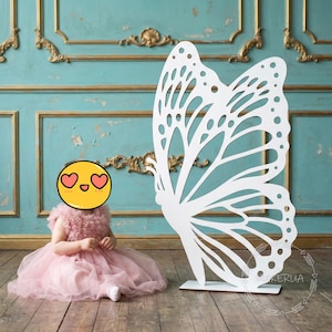 May include: A large, white, butterfly-shaped prop with intricate cut-out details stands on a wooden base. A young child in a pink dress sits on the wooden floor. The background features a blue wall with gold accents.