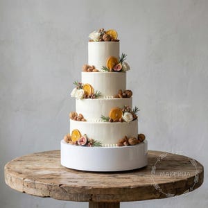 May include: A four-tiered white cake decorated with orange slices, figs, walnuts, and white flowers. The cake sits on a white platform atop a rustic wooden table. The cake is a centerpiece for a celebration.