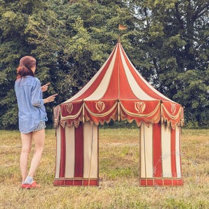 Può includere: Una grande tenda da gioco conica a strisce verticali rosse e crema. La struttura presenta una mantovana smerlata con finiture color oro e motivi ricamati decorativi lungo il bordo superiore, sormontata da una piccola bandiera su un palo centrale.