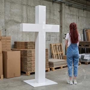 May include: A large, white, cross-shaped sculpture with a glossy finish. The cross stands on a square base. The sculpture is in an industrial setting with cardboard boxes and pallets in the background.