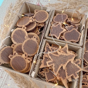 May include: Wooden Christmas ornaments in various shapes, including round, star, and tree designs. The ornaments are displayed in cardboard boxes and feature a perforated pattern. Fairy lights are intertwined among the ornaments.