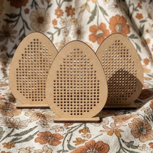 Puede incluir: Tres decoraciones de madera en forma de huevo con numerosos agujeros pequeños. Cada pieza se apoya sobre una pequeña base. La madera es de color marrón claro natural. El fondo es una tela estampada con motivos florales en tonos naranja y crema.
